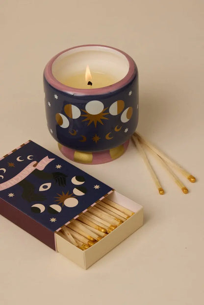 The Jasmine & Rose - A Dopo 8 oz Candle in a celestial-decorated holder sits beside an open box of matches on a beige surface.