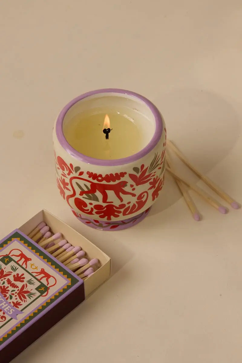 The Coconut & Amber - A Dopo 8 oz Candle in a decorative ceramic holder with red animal and floral designs sits beside three purple-tipped matches and an open matchbox on a beige surface, capturing Mediterranean-inspired style.