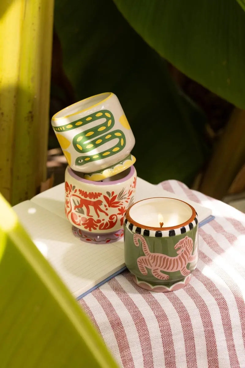 The Coconut & Amber - A Dopo 8 oz Candle in a patterned ceramic holder is displayed on a striped cloth beside an open notebook, with green leaves in the background.