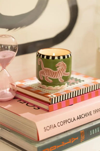 The Black Cedar & Fig - A Dopo 8 oz Candle in a green holder with a tiger design sits on stacked books, including SOFIA COPPOLA ARCHIVE 1999, next to a pink hourglass.