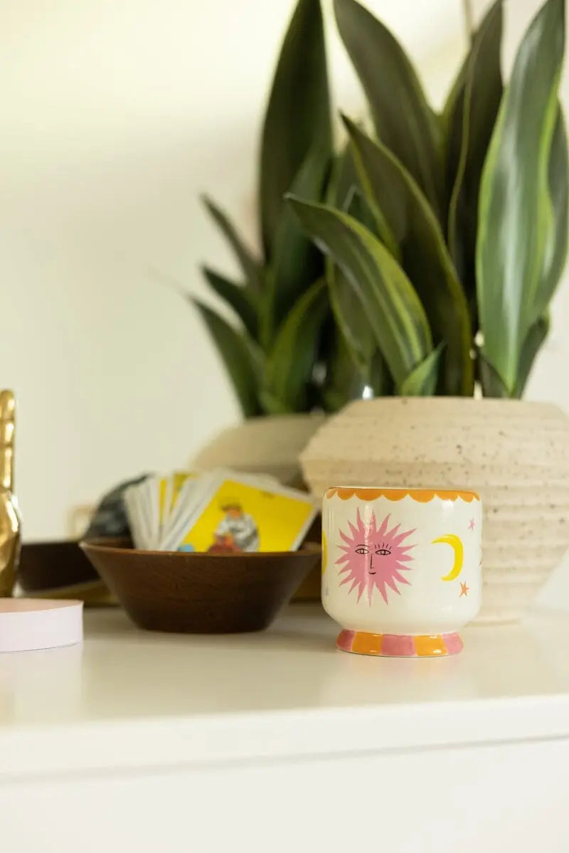 A white tabletop with a decorative sun and moon cup, a bowl of tarot cards, two green plants in beige pots, and the Orange Blossom - A Dopo 8 oz Candle hand-painted to add charm to the serene setting.