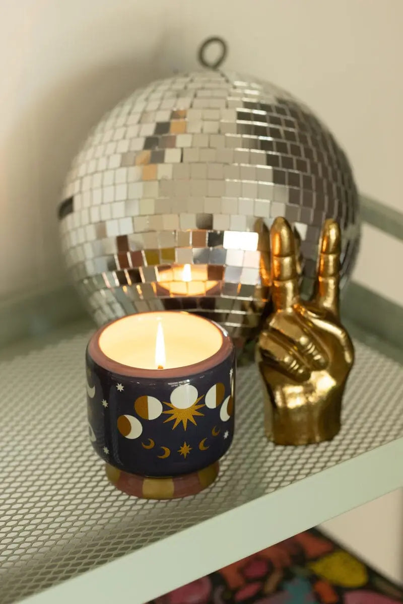 A Jasmine & Rose - A Dopo 8 oz Candle in a moon and sun-themed holder sits beside a gold hand sculpture and mirrored disco ball on a metal shelf.