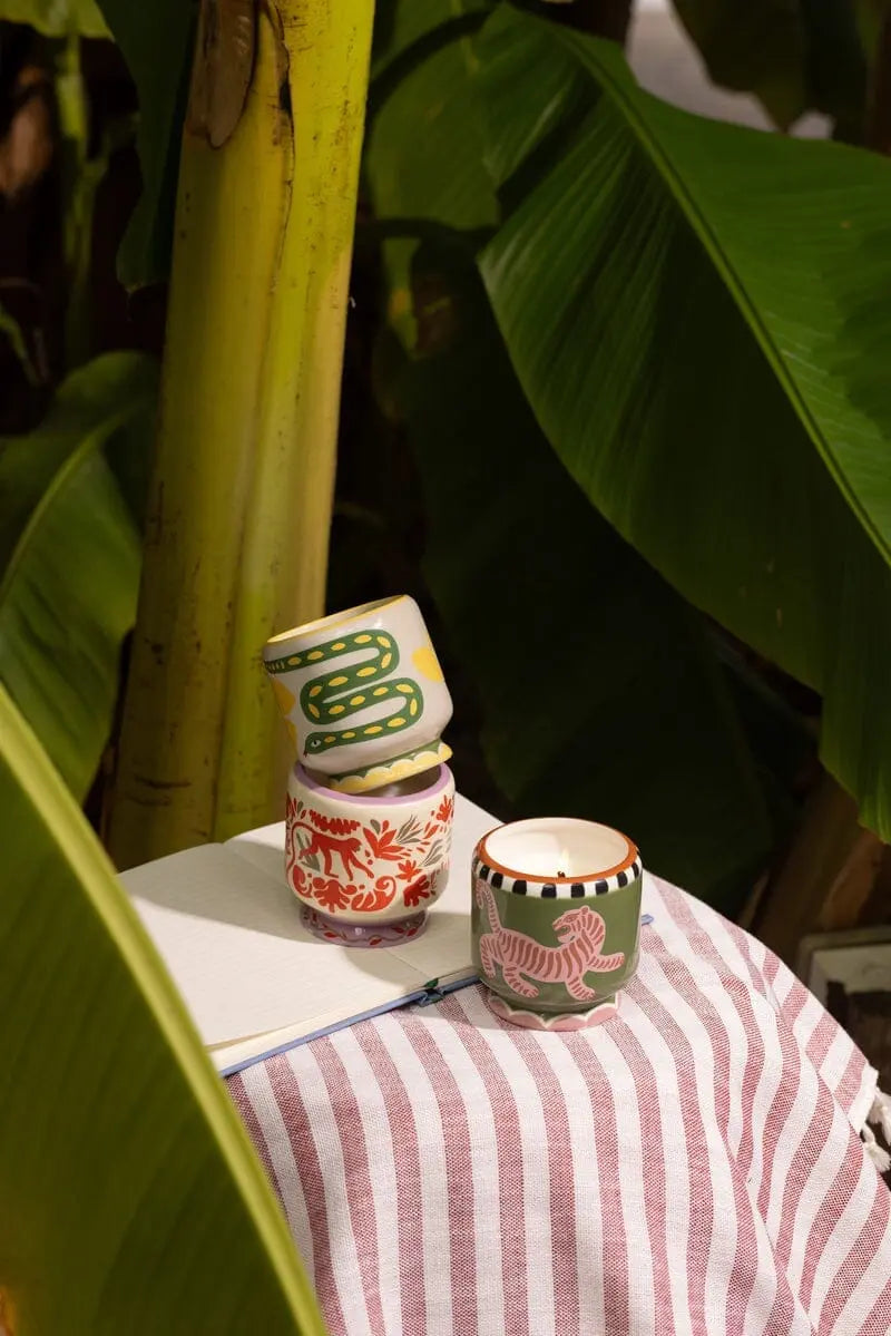 The Wild Lemongrass - A Dopo 8 oz Candle sits on a notebook atop a pink-and-white striped cloth, surrounded by lush green leaves—bringing the fresh charm of Mediterranean fragrances to your space.