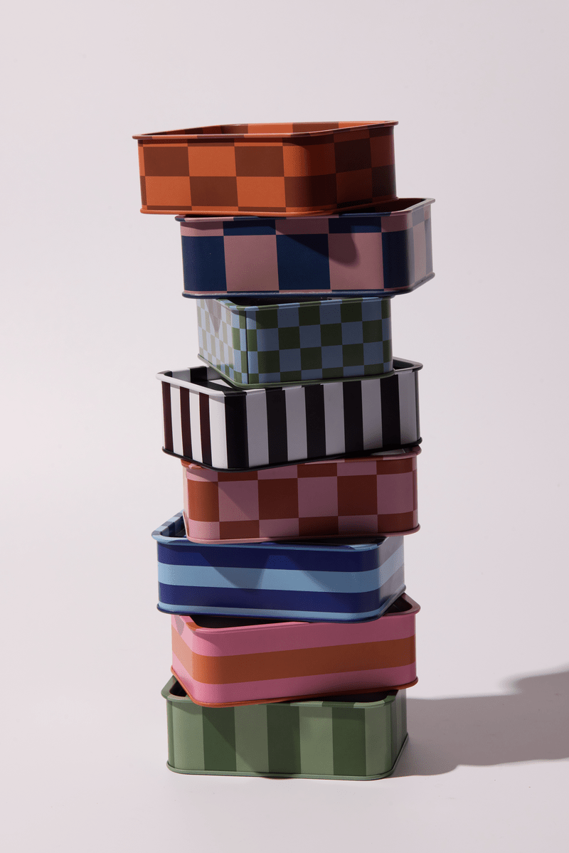 A vibrant stack of seven Cactus Flower - A Dopo 6.5 oz Tin Candles in assorted colors and checkered patterns is arranged on a light surface, casting soft shadows that evoke Mediterranean journeys and the candle’s bold fragrance.