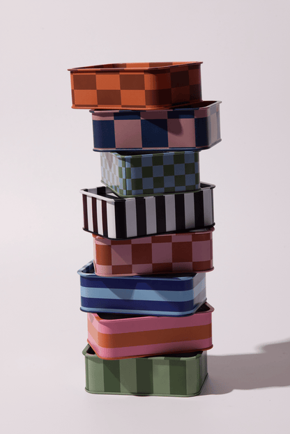 A vibrant stack of seven Cactus Flower - A Dopo 6.5 oz Tin Candles in assorted colors and checkered patterns is arranged on a light surface, casting soft shadows that evoke Mediterranean journeys and the candle’s bold fragrance.
