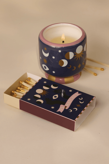 An A Dopo candle in a decorative holder with celestial designs sits beside an open box of A Dopo "Moon" 75ct matches, both featuring matching moon and star motifs inspired by Mediterranean journeys.