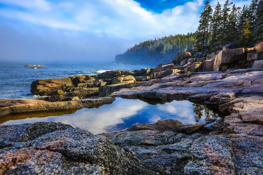 Rocky coastline with rugged cliffs and evergreen trees along the shore. A tide pool in the foreground reflects the blue sky and clouds, while ocean waves crash against the rocks under a partly cloudy sky.