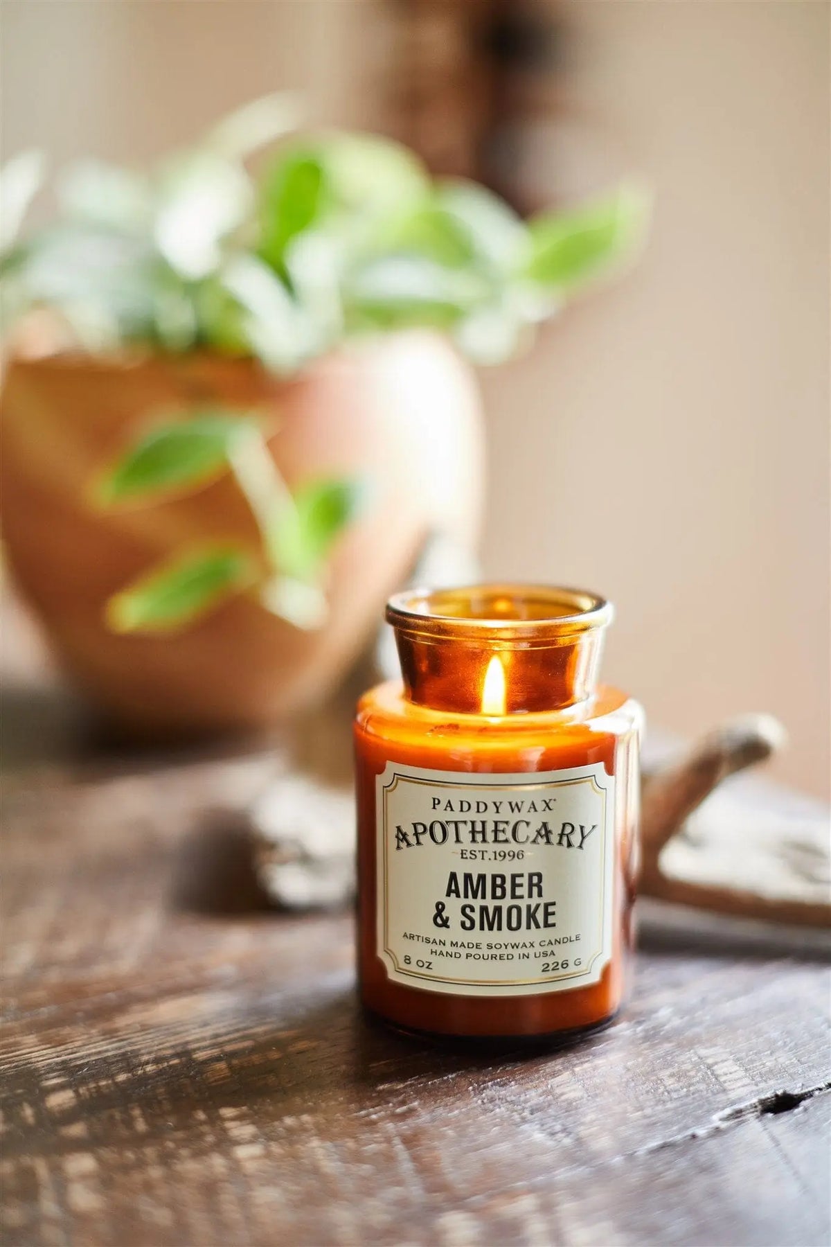 An Amber + Smoke - Apothecary 8 oz Candle burns on a wooden surface, releasing a luxurious fragrance; a potted plant in the blurred background enhances the vintage apothecary vibe.