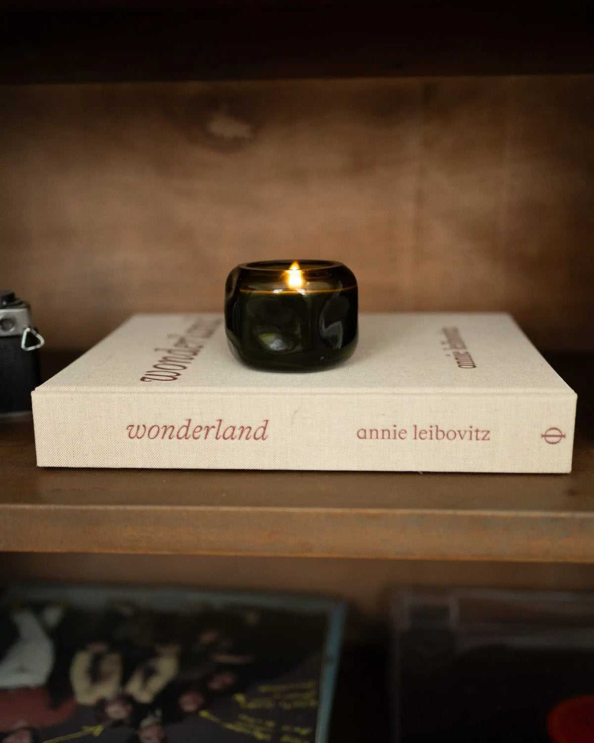 A Basalt 5 oz Wild Fig & Cedar candle, featuring an earthy-woody fragrance, rests in a dark green glass vessel atop the book Wonderland by Annie Leibovitz on a wooden shelf.