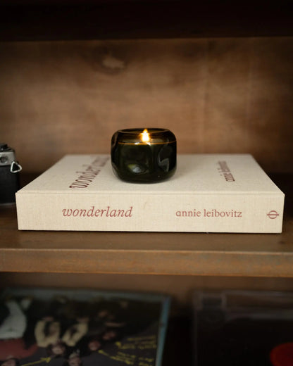 A Basalt 5 oz Wild Fig & Cedar candle, featuring an earthy-woody fragrance, rests in a dark green glass vessel atop the book Wonderland by Annie Leibovitz on a wooden shelf.