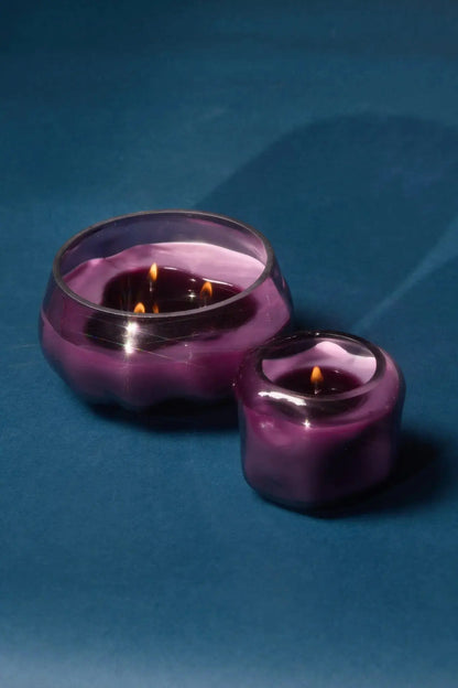 Two purple glass candles with lit flames rest on a blue surface. The larger candle, the Violet & Vanilla - Basalt 10 oz Candle, is housed in an amethyst glass jar and contains three wicks, while the smaller scented candle has a single wick. Their shadows fall to the right side.