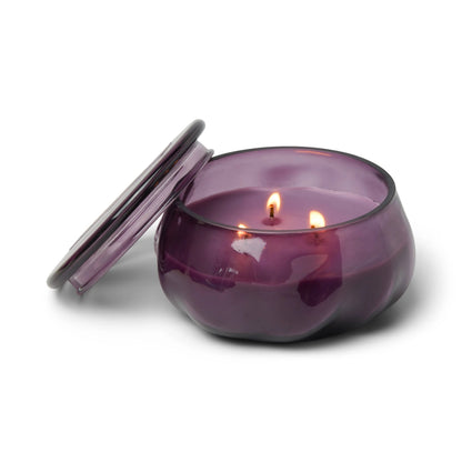 The Violet & Vanilla - Basalt 10 oz Candle glows warmly from its amethyst glass jar, with two wicks gently flickering as the matching lid rests beside it.