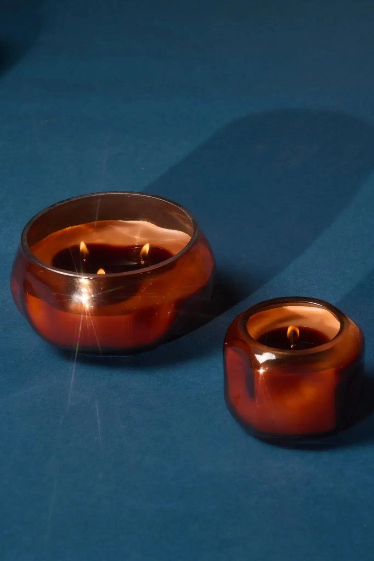 Two copper-colored glass candles with visible flames rest on a blue surface. The larger candle, the Sienna Sunset - Basalt 5 oz Candle, has two wicks, while the smaller features a single wick in an orange-rust glass holder. Both cast soft shadows to the right.