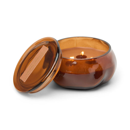 A Sienna Sunset - Basalt 10 oz Candle glows warmly from its brown glass jar, gently releasing its amber fragrance into the air. The lit candle is displayed on a white background with its matching lid resting beside it.