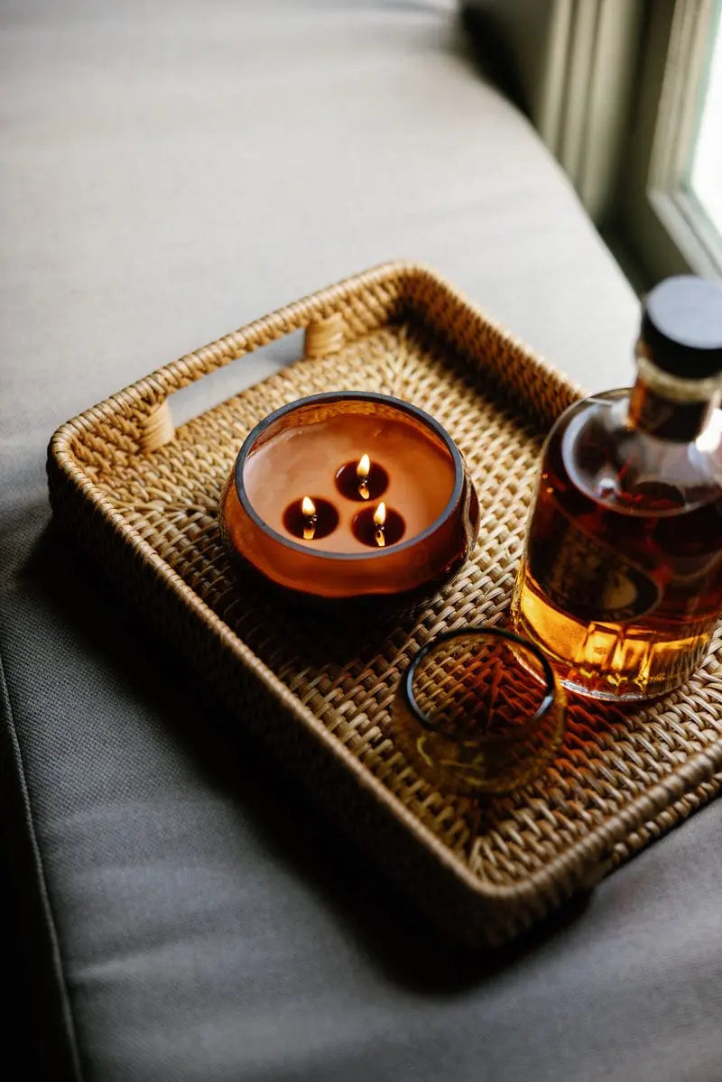 A Sienna Sunset - Basalt 10 oz Candle with three flames, a glass of amber liquid, and a bottle rest on a woven tray atop a gray couch near a window, filling the space with its warm amber fragrance.