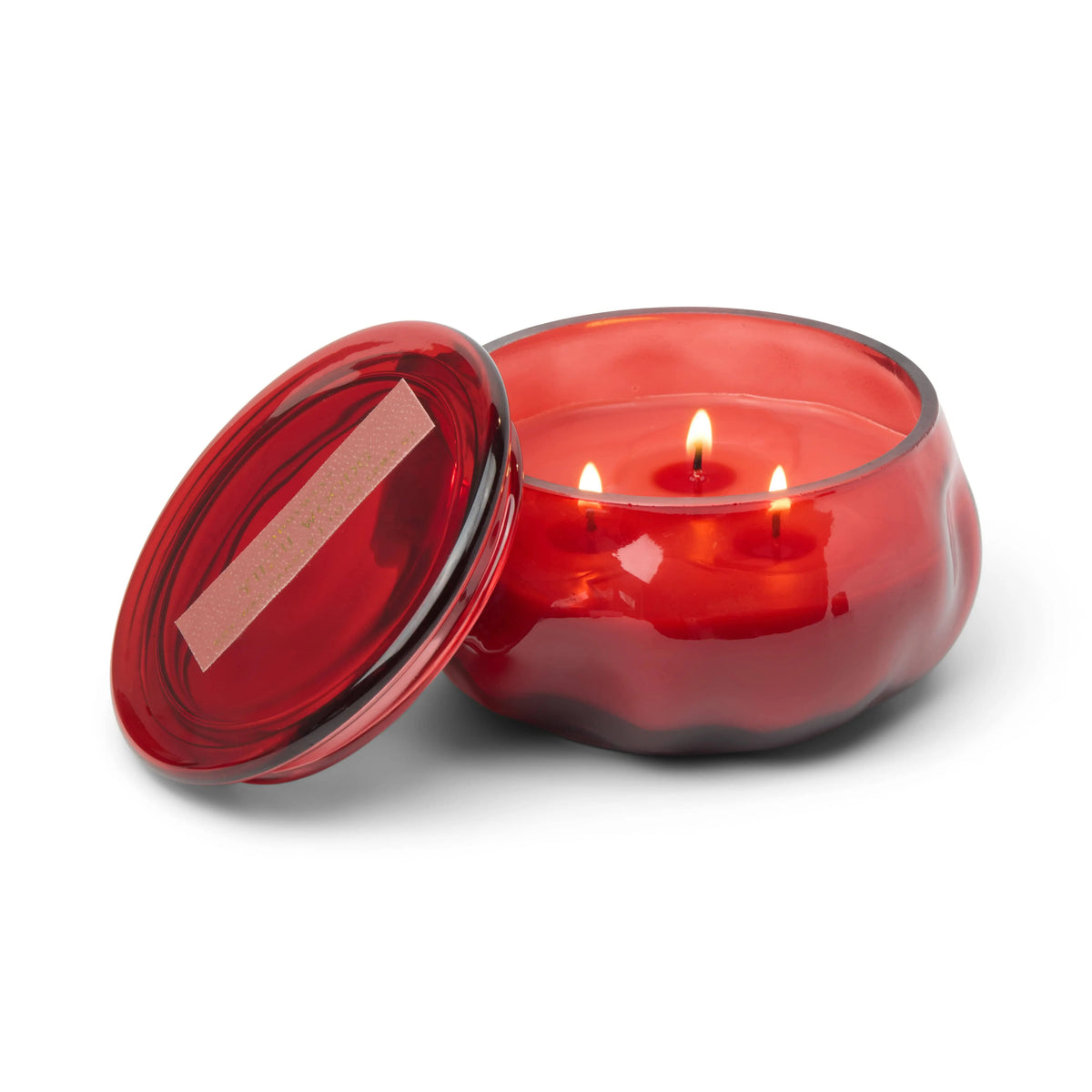 A wavy red glass jar sits open with the lid beside it, holding the Yuzu Woods - Basalt 10 oz Candle inside. Three lit wicks fill the scene with a warm citrus glow.