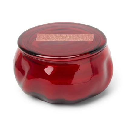 A round, deep red glass jar with a matching lid and a gold label on top, the Yuzu Woods - Basalt 10 oz Candle is strikingly displayed against a white background.