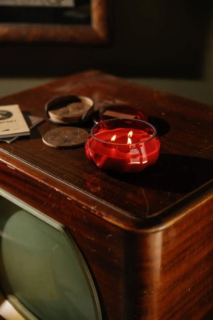 A red glass ashtray holding a lit Yuzu Woods - Basalt 10 oz Candle sits atop a vintage wooden TV, accompanied by coasters and scattered papers, creating a nostalgic, warm atmosphere enriched with subtle citrus notes.