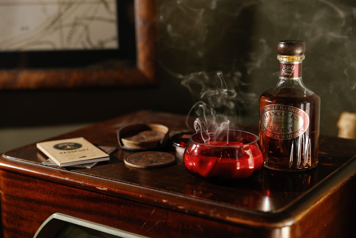 A bottle of whiskey, a red ashtray with smoke rising, a closed cigar case, cigarettes, and a coaster sit on a polished wooden table near a framed picture.