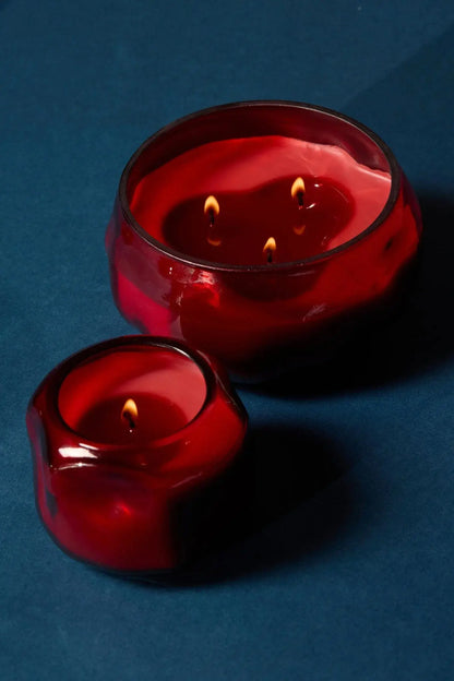 Two red glass candles rest on a dark blue surface. The larger candle—the Yuzu Woods - Basalt 10 oz Candle—glows with three flames, while the smaller one in a coordinating red glass jar has a single flame. Both citrus-scented candles are lit, their rounded, reflective surfaces catching the light.