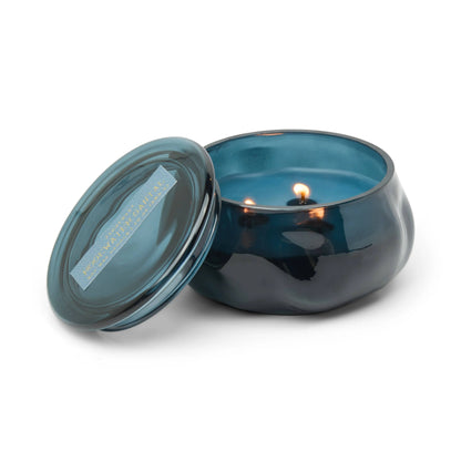 A blue metallic tin candle, featuring a subtly wavy texture and glowing with two flames within, sits open beside its matching lid marked “Noble Platinum.” This floral-scented Rosewater Santal - Basalt 10 oz Candle evokes elegance and tranquility, its shimmering blue glass jar catching the light and infusing any space with refined charm.