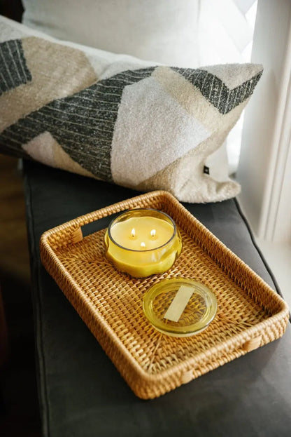 A Tobacco Patchouli - Basalt 10 oz Candle in a yellow glass jar with the lid off rests on a woven tray atop a black cushioned bench near a geometric-patterned pillow, filling the space with its warm fragrance.