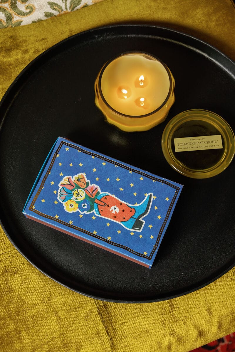 A lit candle, a small jar labeled TOBACCO PATCHOULI, and Cowboy Boot - Large Rectangle Boxed Matches sit on a black tray atop a yellow fabric surface.
