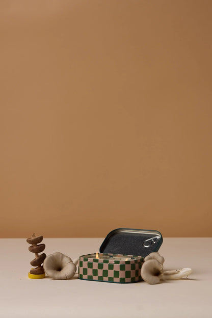 An open Wild Mushroom - Bistro 4.5 oz Tin Candle rests on a green-and-white checkered surface, surrounded by light mushrooms and set against a beige background, capturing the cozy charm of the Bistro Collection.