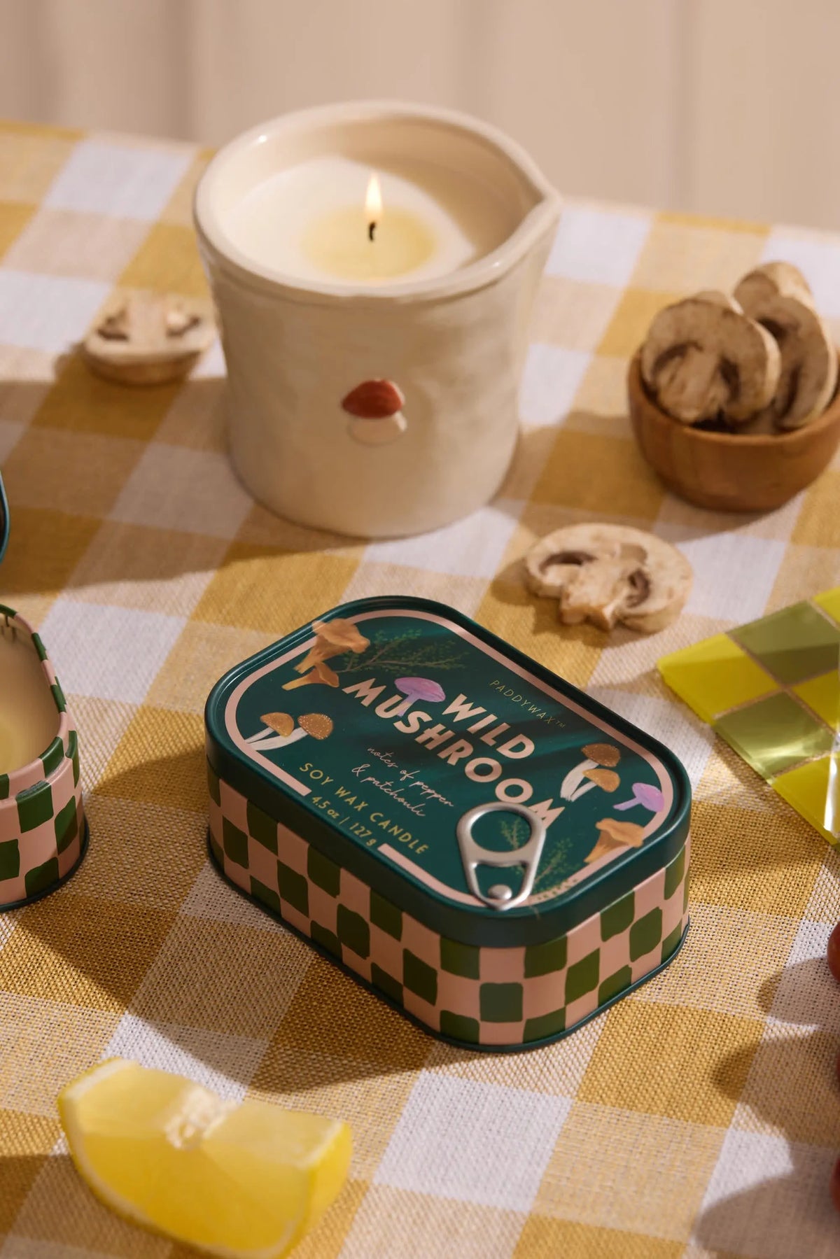 A Wild Mushroom - Bistro 4.5 oz Tin Candle rests on a yellow-checkered tablecloth, surrounded by dried mushroom slices, a lit ceramic candle, and a yellow-green checkered matchbox.
