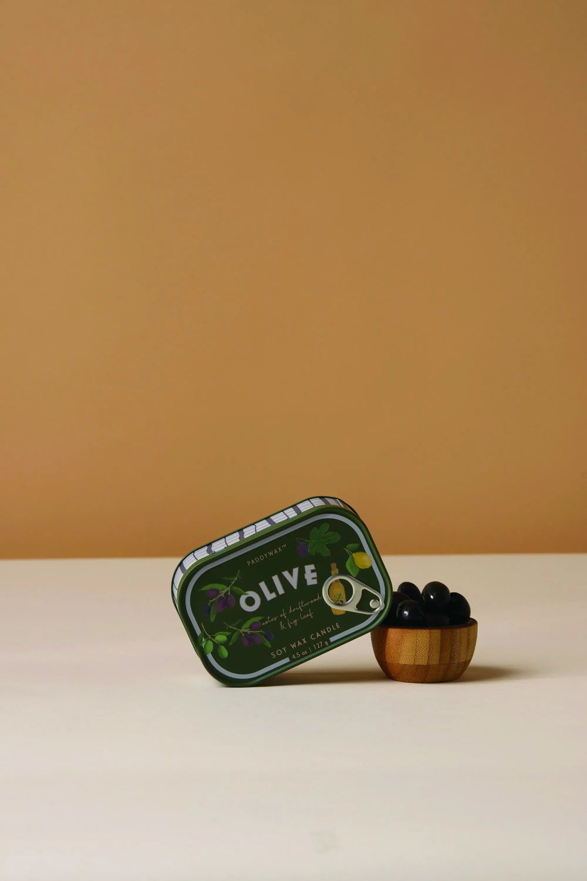 A green tin labeled Fig & Olive - Bistro 4.5 oz Tin Candle leans against a small wooden bowl filled with black olives, set on a light surface with a plain beige background.