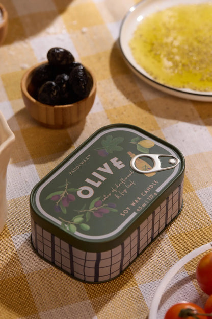 A tin from the Bistro Collection labeled Fig & Olive - Bistro 4.5 oz Tin Candle sits on a checkered tablecloth, surrounded by a small wooden bowl of black olives, a plate with olive oil, and some tomatoes. This beautifully designed candle brings the ambiance of a Mediterranean bistro right to your table.