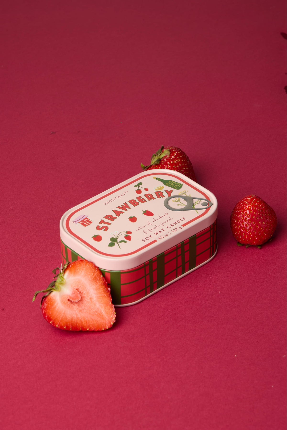 The Strawberry - Bistro 4.5 oz Tin Candle, featuring a strawberry design and label, rests on a red surface with two whole strawberries and one halved strawberry nearby.