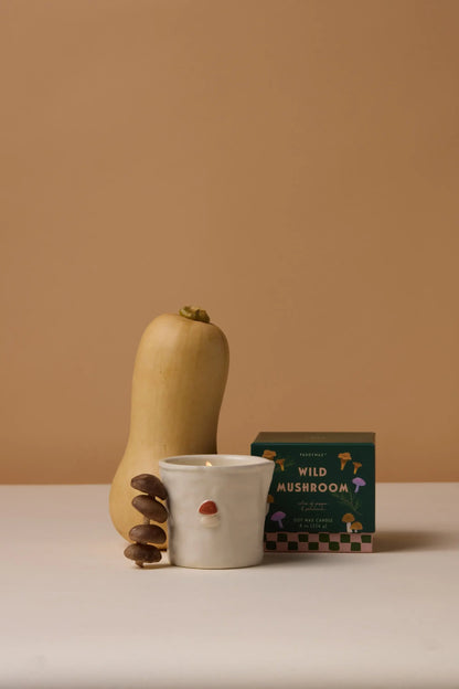 A butternut squash, mushrooms, an aromatic Wild Mushroom - Bistro 8 oz Candle in a ceramic cup with a small red dot, and a green box are arranged on a white surface against a beige background.