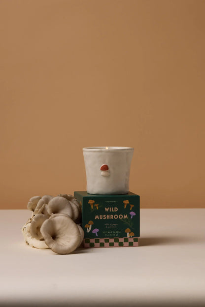 The Wild Mushroom - Bistro 8 oz Candle, a white ceramic candle with a small nose detail, sits on its green box beside oyster mushrooms—a delightful gift for candle lovers.