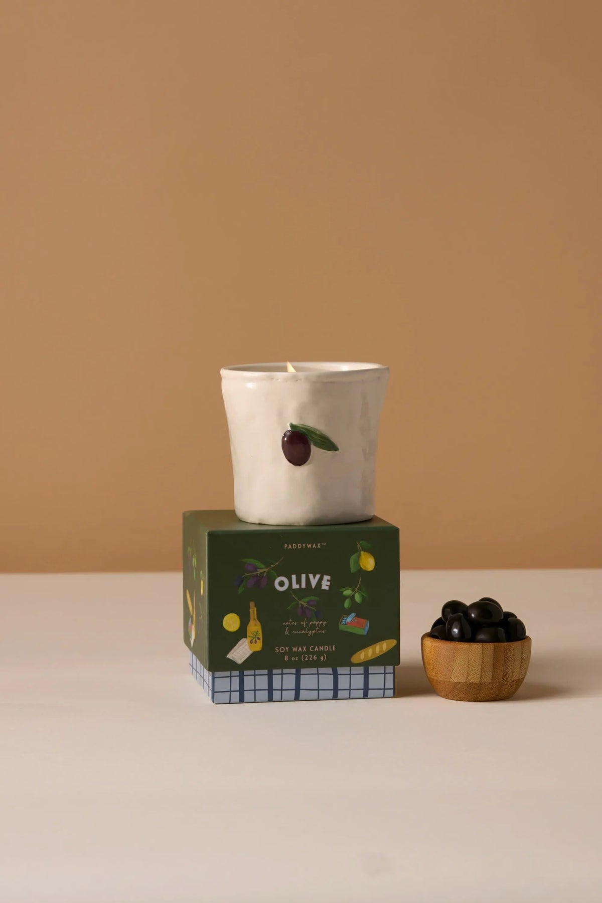 The Fig & Olive - Bistro Candle 8 oz, a white ceramic candle with hand-painted food and olive designs, rests atop its green box beside a wooden bowl of black olives.