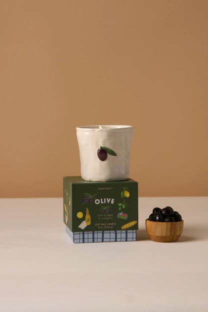 The Fig & Olive - Bistro Candle 8 oz, a white ceramic candle with hand-painted food and olive designs, rests atop its green box beside a wooden bowl of black olives.