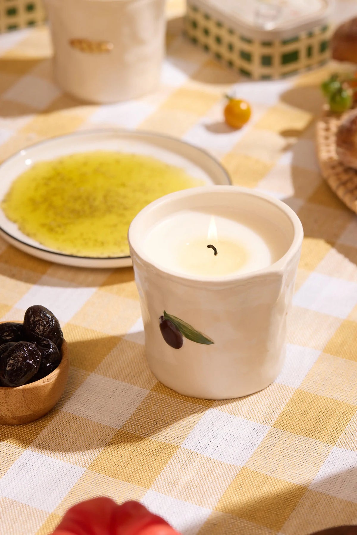 The Fig & Olive - Bistro Candle 8 oz, a white ceramic holder with a hand-painted olive design, sits on a checkered tablecloth with ceramic candles and olive oil, evoking the cozy charm of a Parisian café.