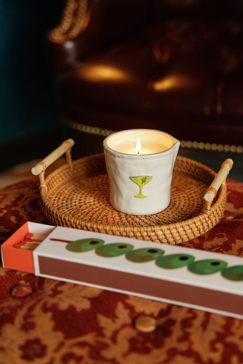 A lit candle in a white cup with a martini glass design sits on a woven tray beside Olives - Extra Long Boxed Matches, all arranged on a red patterned surface with a leather chair in the background.