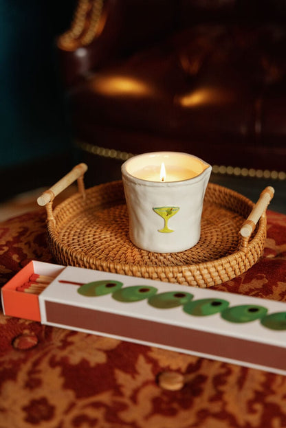 A lit candle in a white cup with a martini glass design sits on a woven tray beside Olives - Extra Long Boxed Matches, all arranged on a red patterned surface with a leather chair in the background.