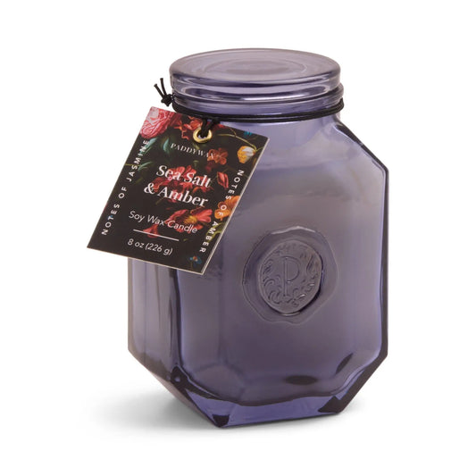 The Sea Salt + Amber - Botanica 8 oz Candle features purple glass, a black lid, and a floral tag—an elegant choice for home fragrance from the luxurious Botanica collection.