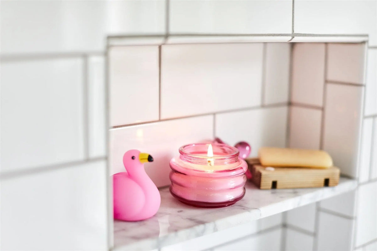 A pink flamingo figurine, the Desert Peach - Beam 3 oz Candle casting ambient light in a glass jar, and a soap bar on a dish rest on a marble shelf set into a white tiled wall.
