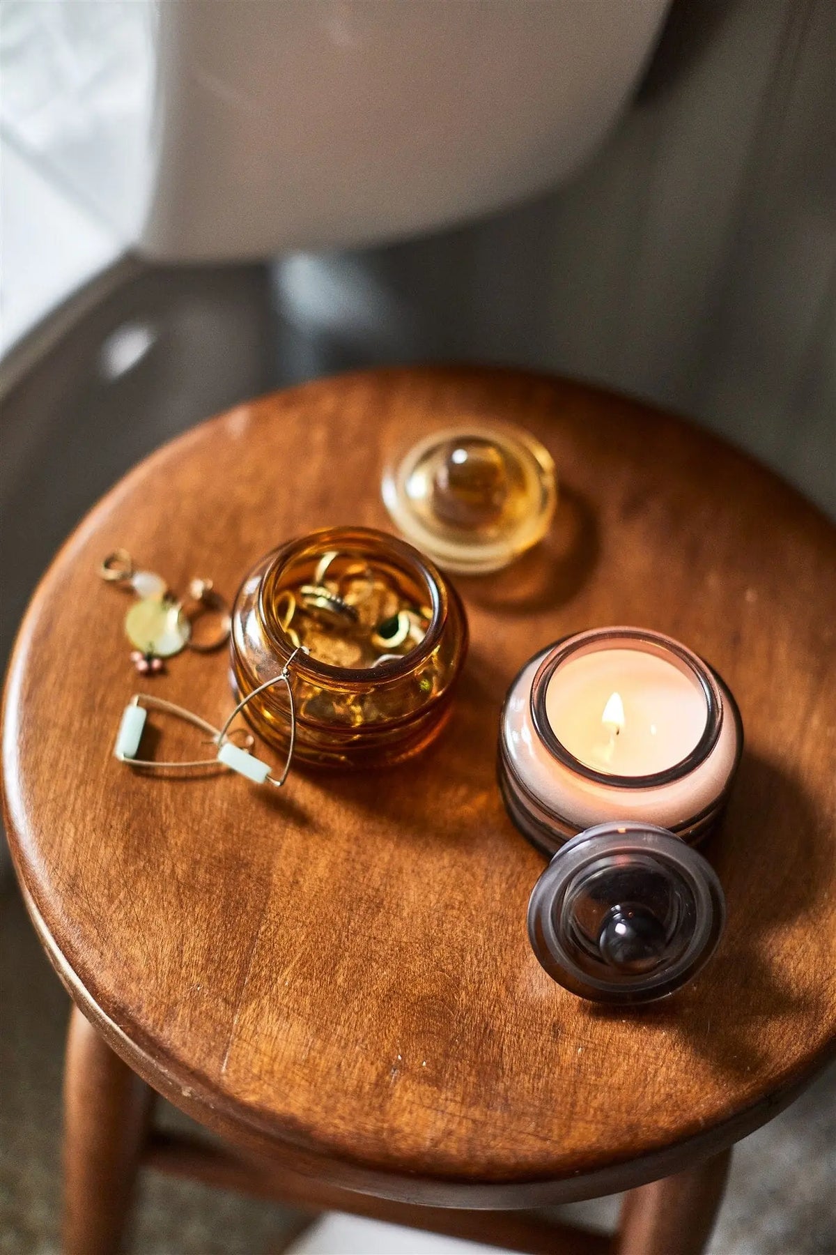 A wooden stool holds a jar of capsules, gold earrings, and the Baltic Ember - Beam 3 oz Candle, which fills the space with a luxurious fragrance and warm ambient light.