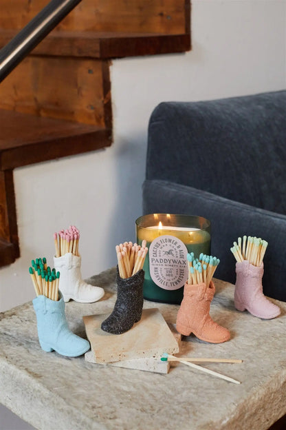 A black ceramic Cowboy Boot Match Holder filled with matches sits on a stone table beside a lit candle, with a staircase and sofa in the background, enhancing the Western atmosphere.