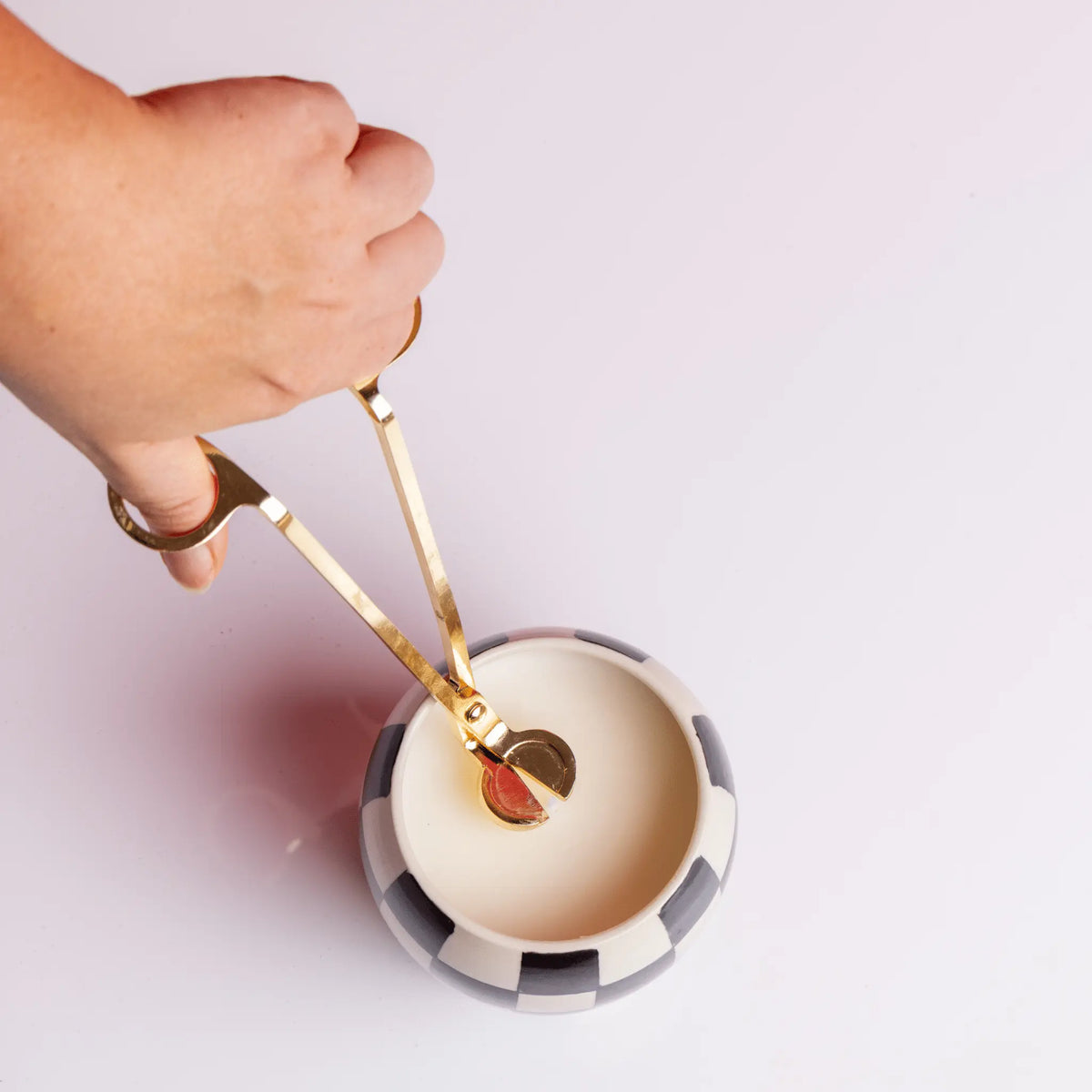 A hand uses a Wick Trimmer to cut the wick of a white candle in a striped holder on a white surface.