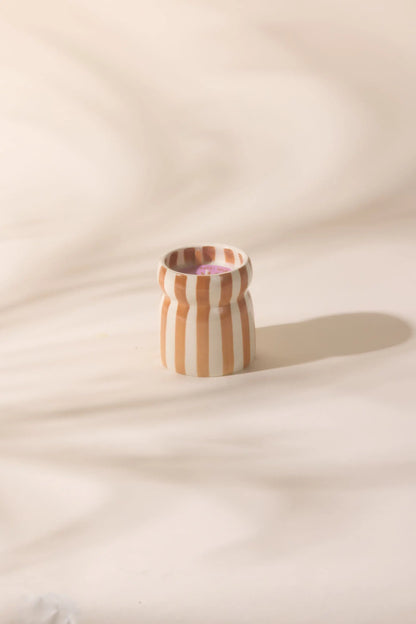 The Sienna Sunset - Cabana 6.5 oz Candle sits in a small ceramic vessel with vertical brown and white stripes on a softly shadowed surface, exuding coastal scents and artisanal charm.