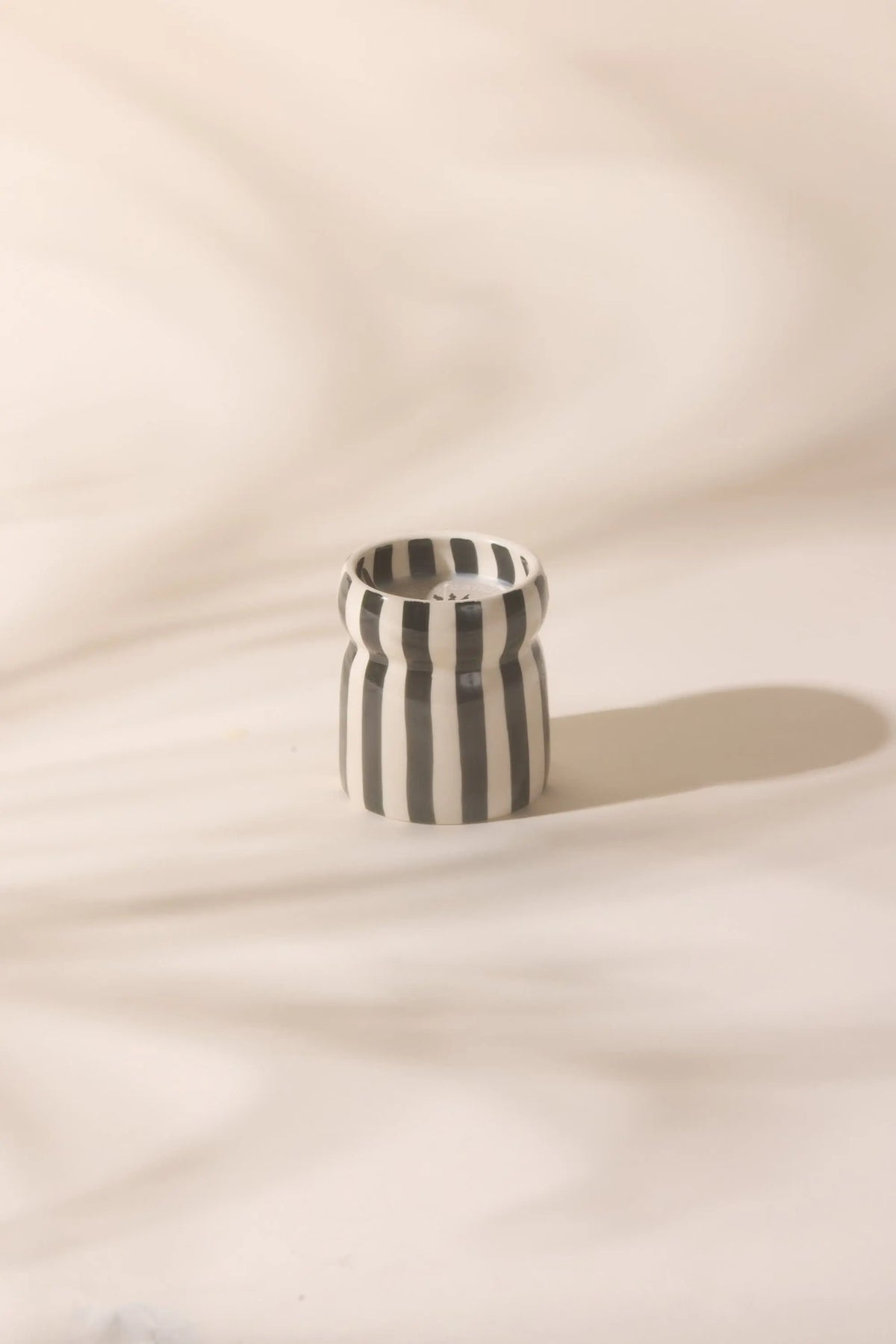 The Lava Stone - Cabana 6.5 oz Black Candle, a small ceramic vessel with black and white vertical stripes, sits on a light surface with soft shadows in the background.