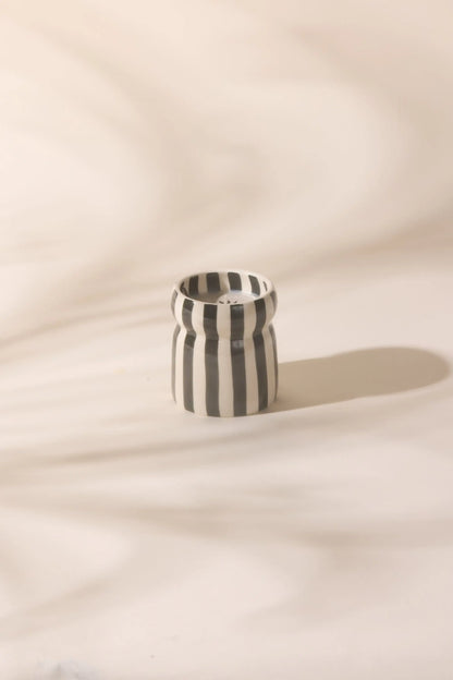 The Lava Stone - Cabana 6.5 oz Black Candle, a small ceramic vessel with black and white vertical stripes, sits on a light surface with soft shadows in the background.