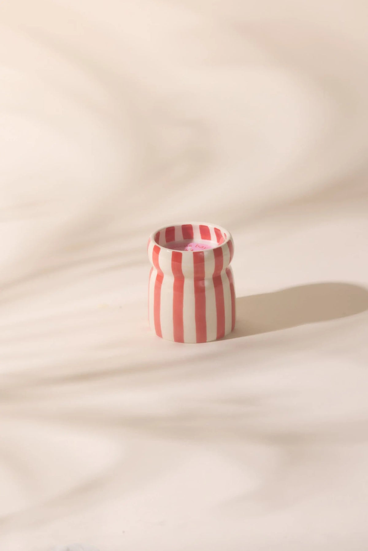 The Hibiscus Bay - Cabana 6.5 oz Candle, a small ceramic vessel with vertical red and white stripes, rests on a light surface, evoking coastal scents in gentle, diffused light.