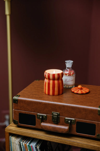A glowing Spiced Apple - Cabana 6.5 oz Candle with red and orange stripes sits atop a brown suitcase beside its lid and a clear glass jar labeled "Matches," filled with red-tipped matches.