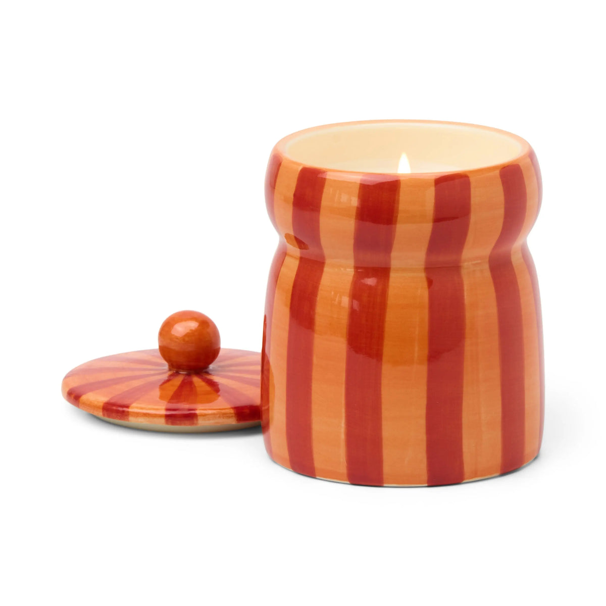 A Spiced Apple - Cabana 6.5 oz Candle burns inside a striped orange and red ceramic jar, with its matching lid placed beside it.